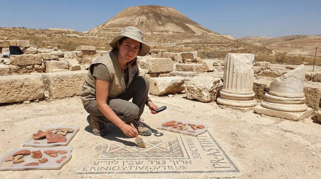 Israeli archaeologist examines mosaic floors at Herodium, artifacts in background.