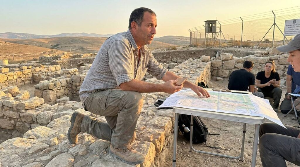 Israeli historian examines artifacts at archaeological site with stone ruins backdrop.