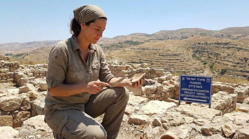 Israeli archaeologist examines ancient ruins in Hamonah, hills in background.